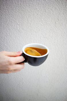 A hand holding a black cup of aromatic espresso coffee against a neutral background.