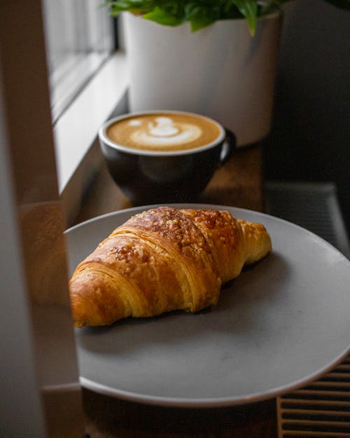 Plate with baked sweet croissant and cup of delicious cappuccino placed on windowsill in daylight
