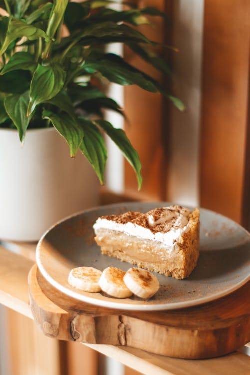 Delicious freshly baked cake on plate decorated with ripe banana and served on wooden shelf near lush houseplant