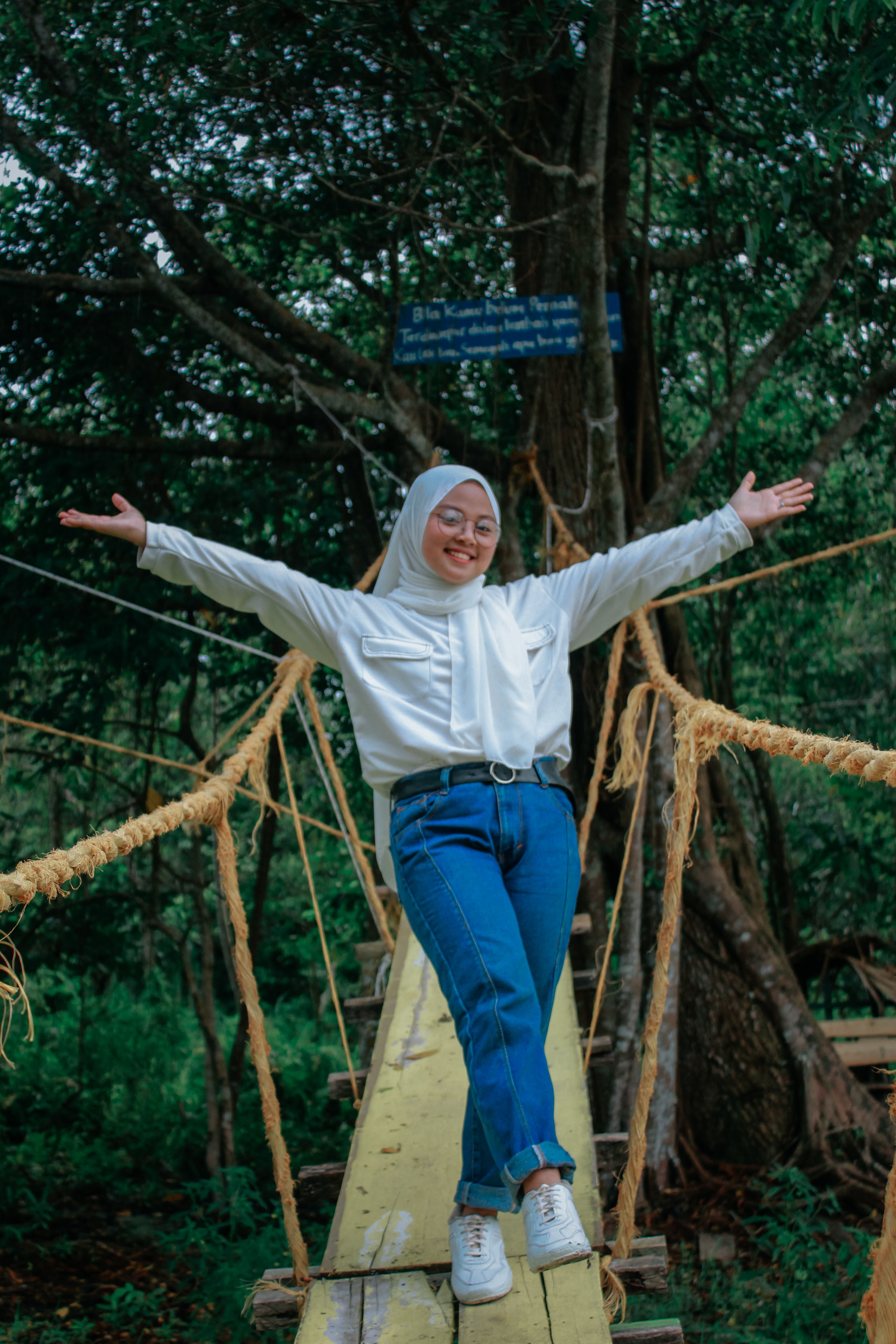 Smiling woman with arms raised standing on a rope bridge in lush green forest, Indonesia.