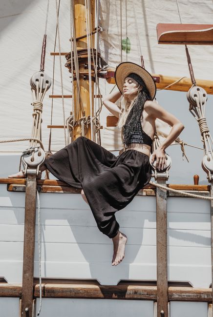 Full body side view of barefoot female in hat sitting on wooden hull of ship during cruise on summer day