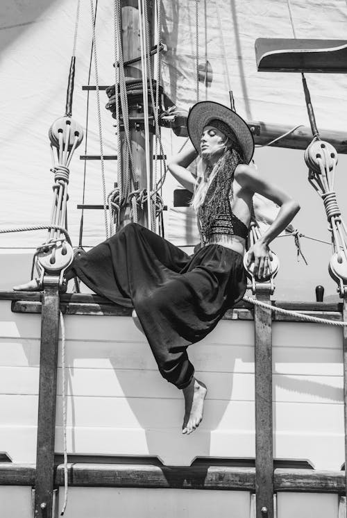 Black and white full body side view of stylish female in hat sitting on wooden ship during cruise on summer day