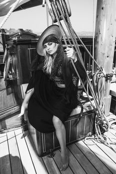 Black and white full body of trendy barefoot female in hat looking at camera while sitting on deck of ship during trip
