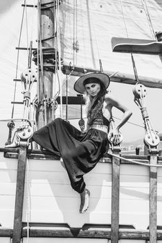 Black and white full body of confident barefoot female in hat looking at camera while sitting on hull of ship