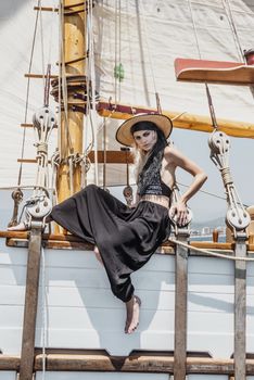 Full body of barefoot female in stylish outfit and hat looking at camera while sitting on wooden hull of ship during trip