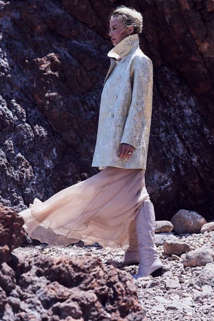 A stylish woman in a textured coat and skirt posed dramatically against rugged rocks.
