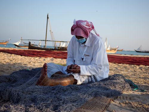 Free Elderly fisherman in traditional attire mends a net on a sandy beach with dhows in the background. Stock Photo