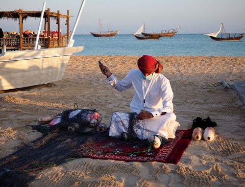 Free Man mends fishing net on sandy shore with boats in background. Stock Photo