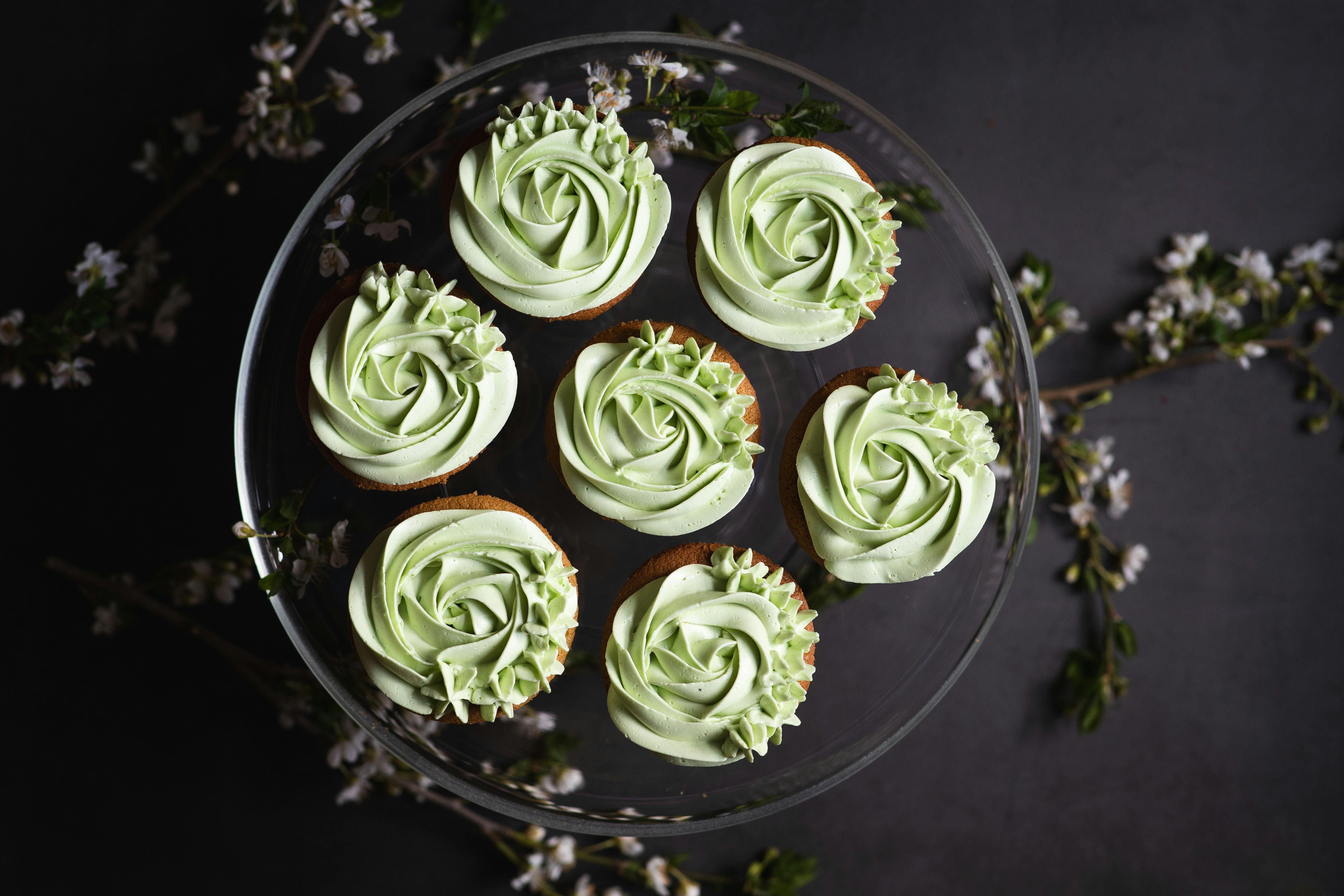 Delicate green frosted cupcakes arranged on a glass stand with floral accents, perfect for celebrations.