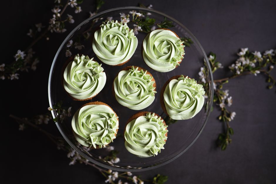 Delicate green frosted cupcakes arranged on a glass stand with floral accents, perfect for celebrations.