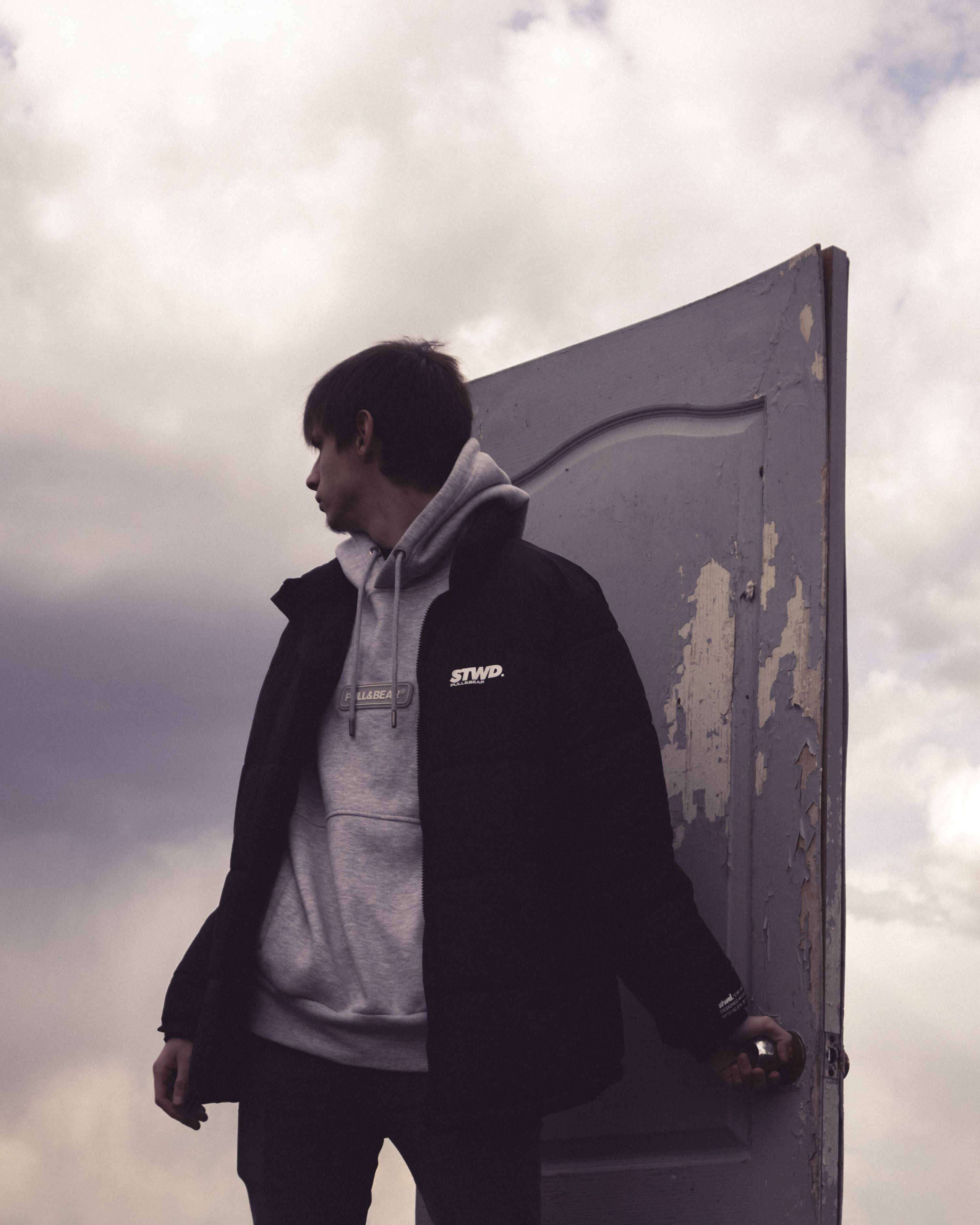 A young man in a hoodie stands by a weathered door, looking back against a cloudy sky.