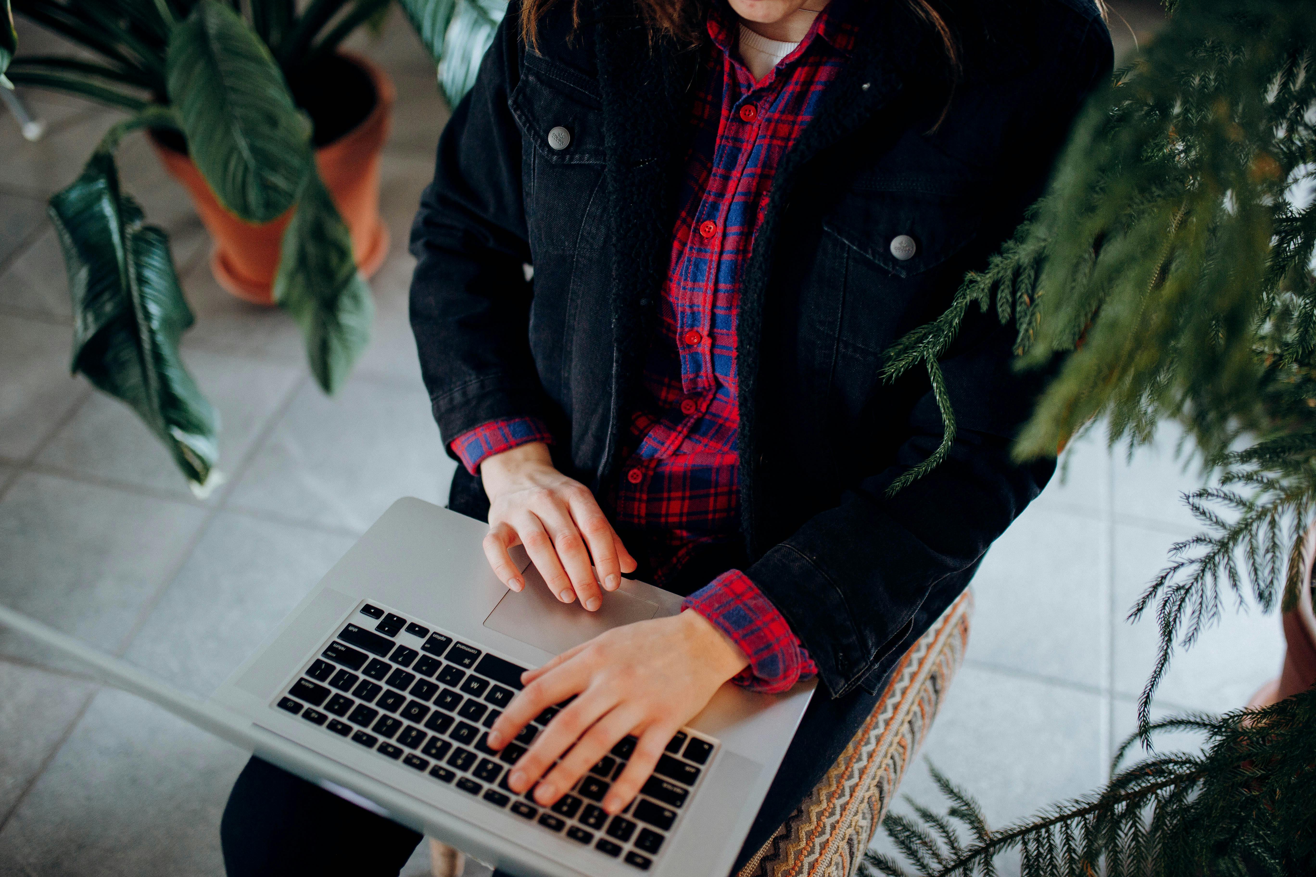 Close-up of hands typing on a laptop amidst houseplants, wearing a denim jacket and plaid shirt.