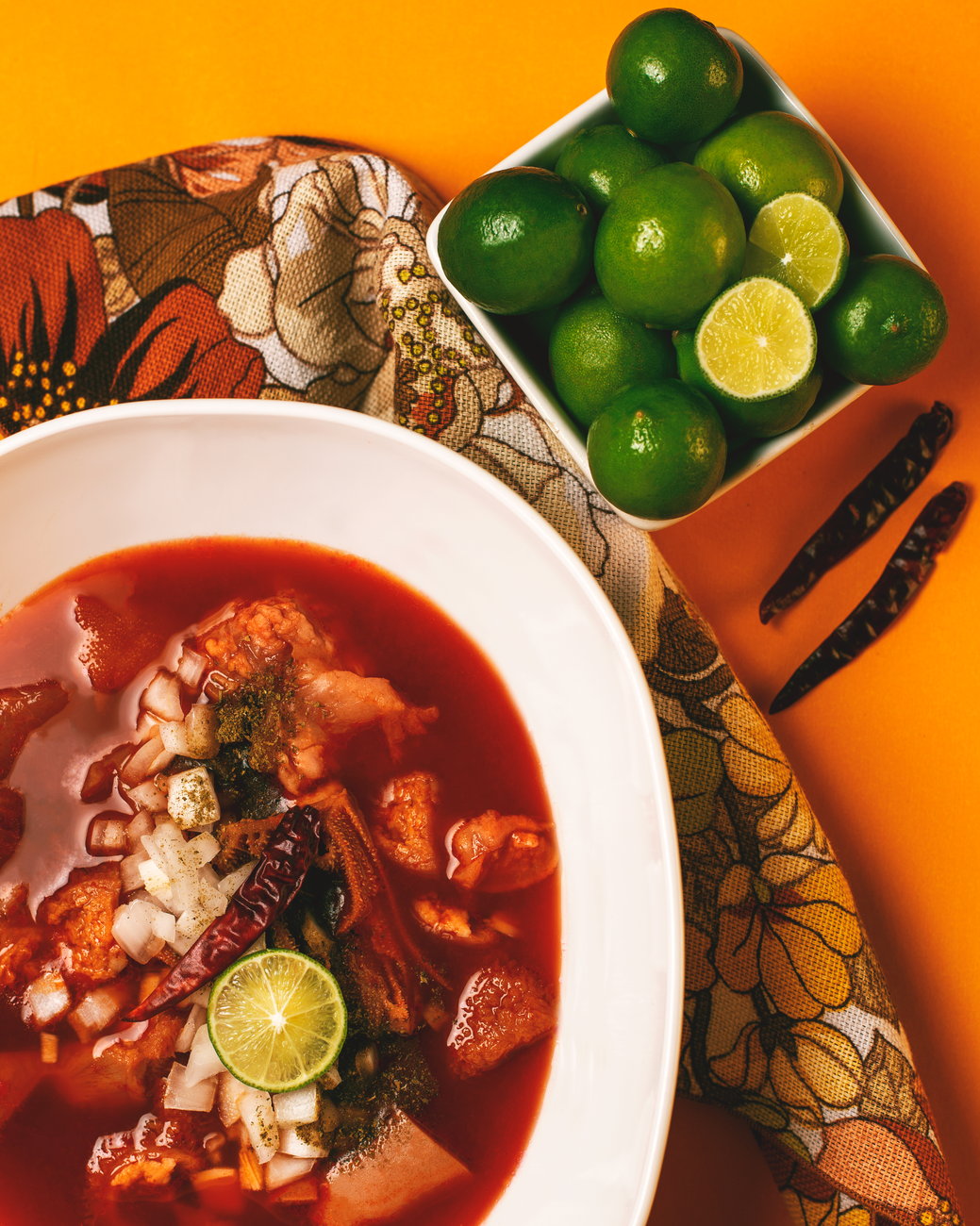 Bowl of red pozole soup with traditional garnishes including lime wedges, sliced radishes, oregano, and chile powder