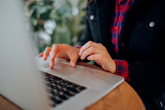 Person wearing a plaid shirt typing on a laptop keyboard indoors, focusing on hands and device.