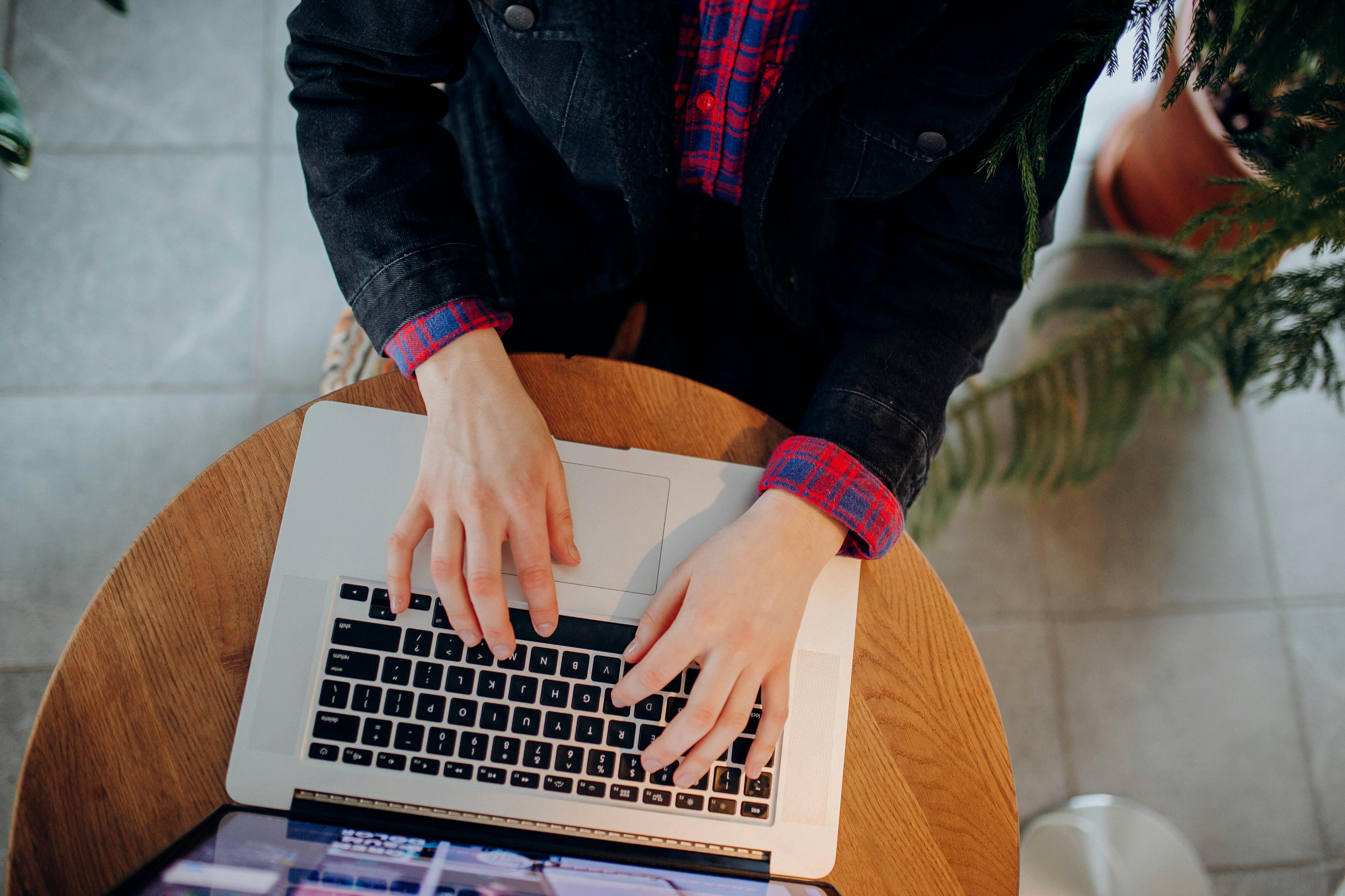 Hands typing on a laptop keyboard from above, surrounded by plants indoors.