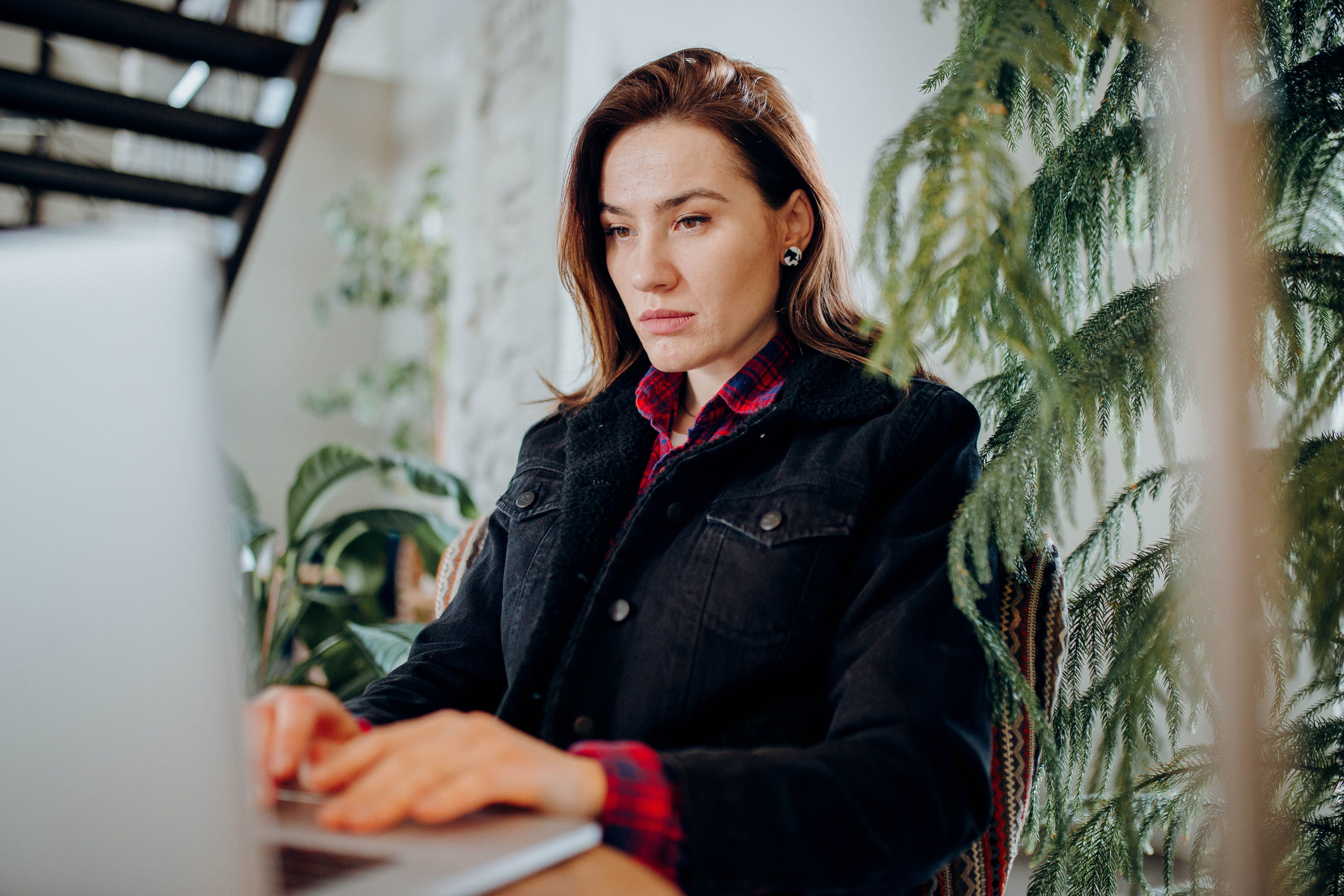 A woman in a denim jacket works intently on her laptop in a plant-filled environment.