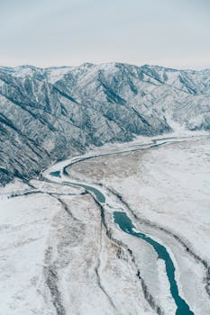 A serene aerial view of a winding river amidst snow-covered mountains in winter.