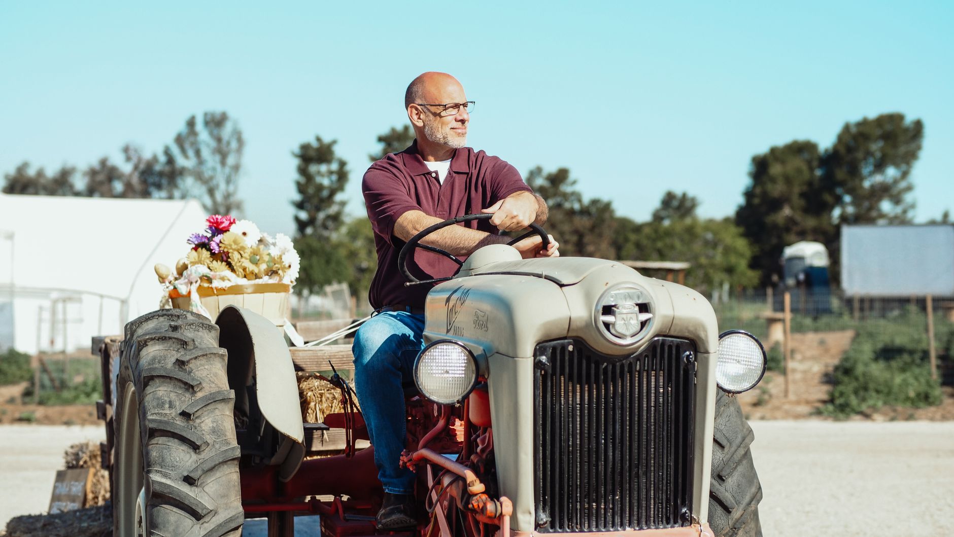 Elderly man operating a vintage tractor in an open farm setting, surrounded by clear skies.
