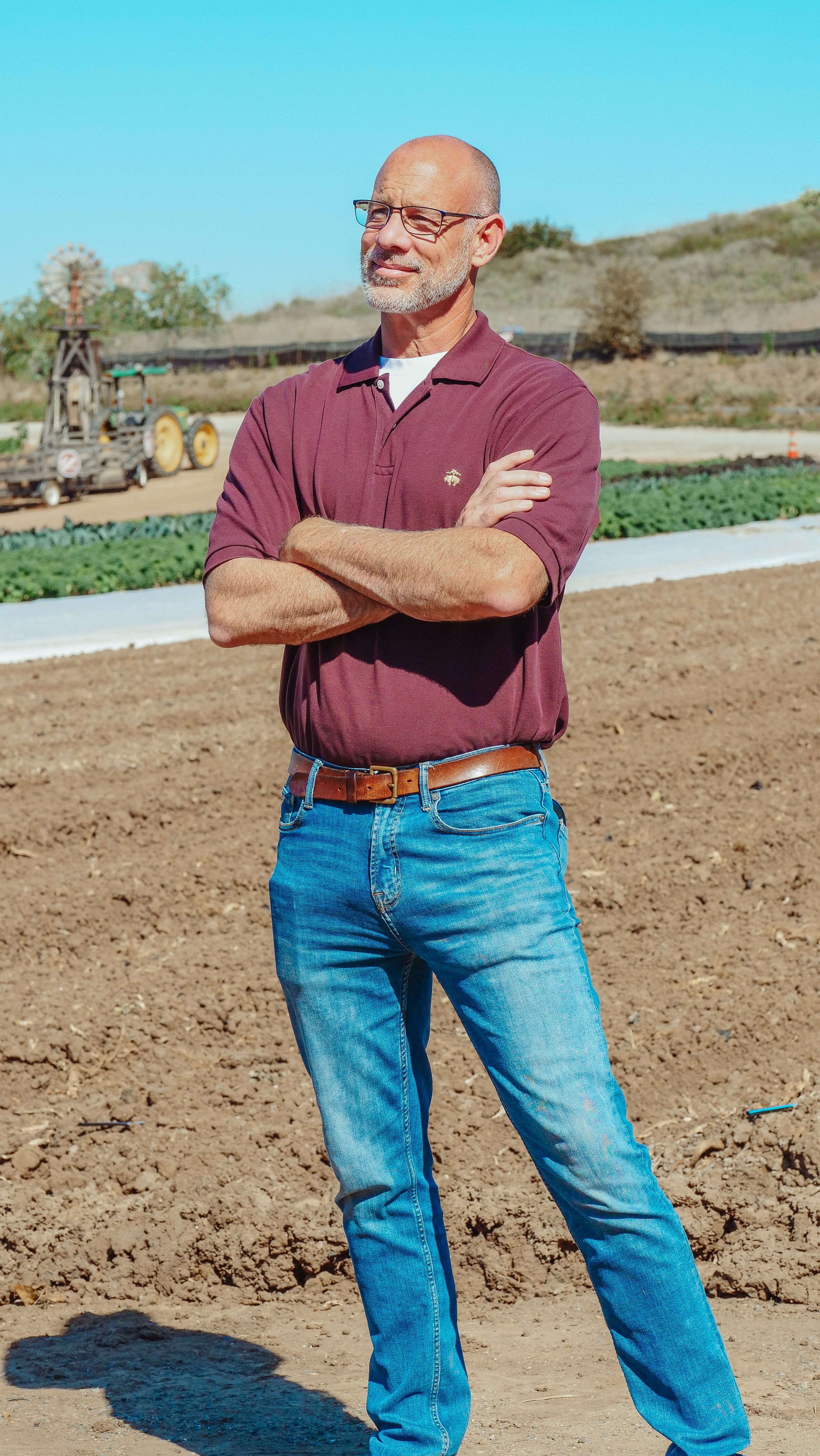 Senior farmer with arms crossed standing in a sunny rural farm setting, showcasing traditional agriculture.
