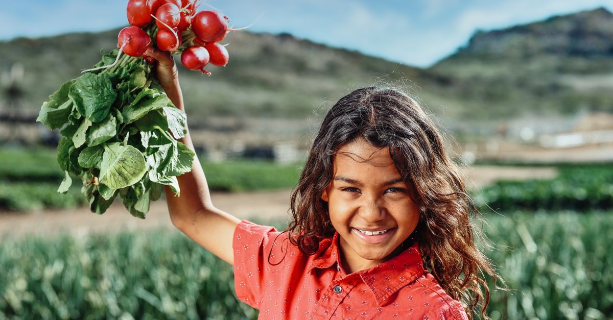 Photo by Kindel Media Smiling girl holding red radishes in a field under a blue sky during daytime.