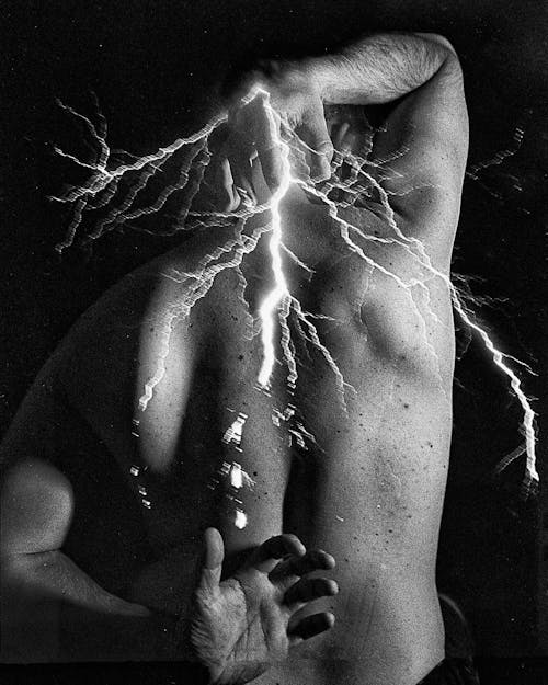 Back view of black and white crop unrecognizable shirtless muscular male standing in darkness with artificial lightning in hands