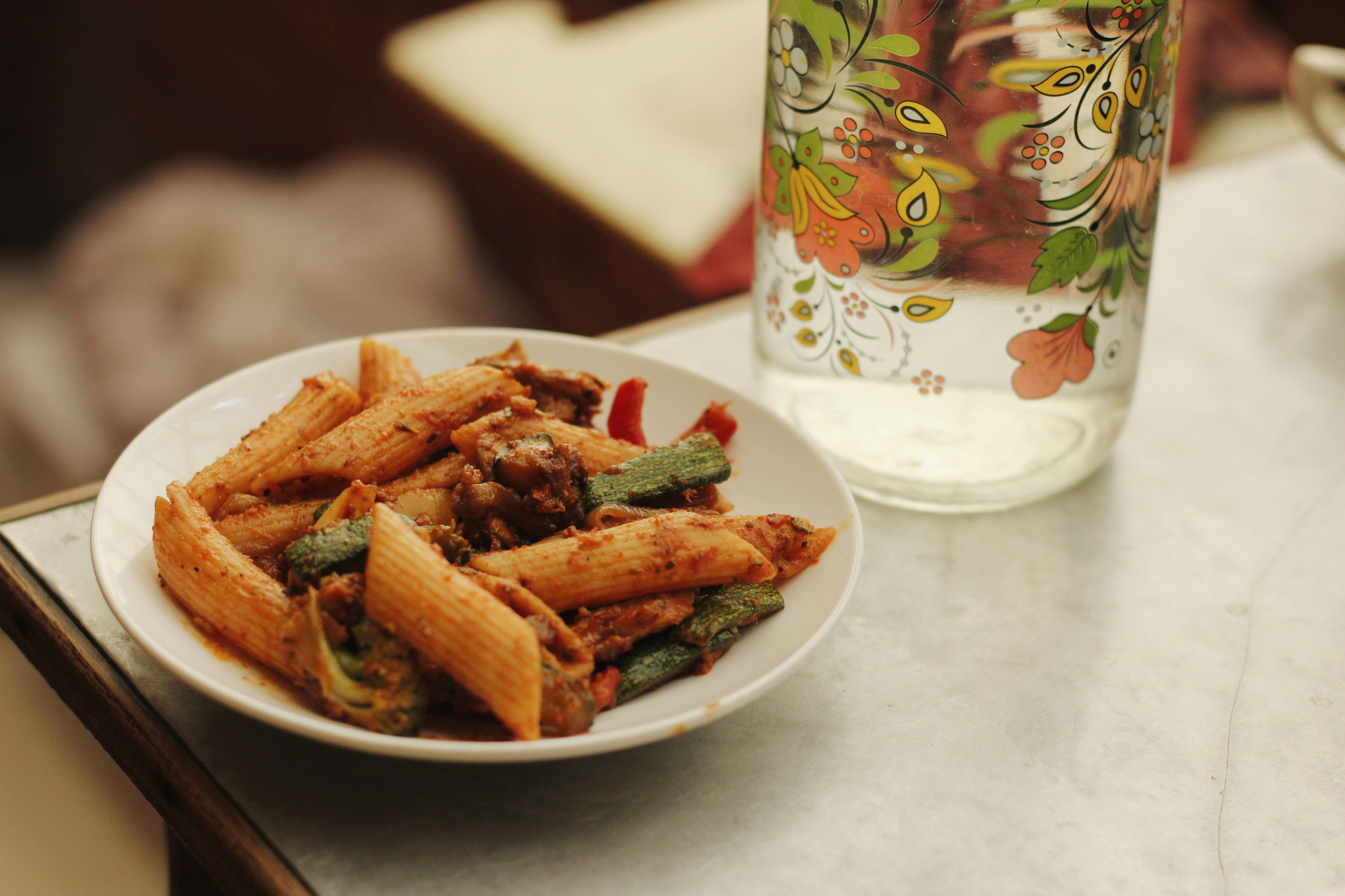 Close-up of penne pasta with vegetables indoors, paired with a colorful floral jug.