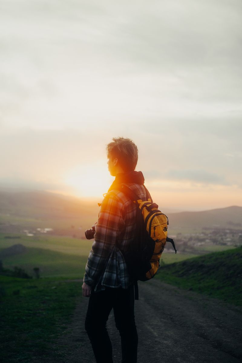 Back view of male hiker with rucksack and photo camera admiring mounts against shiny sunlight at dawn