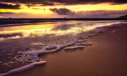 Free Serene beach scene showcasing a stunning sunset with golden reflections and gentle sea foam. Stock Photo