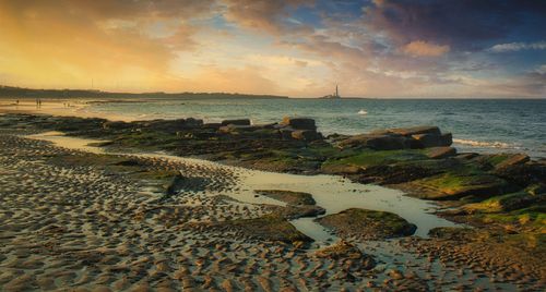 Free A serene seascape with a rocky shoreline at sunset, showcasing dramatic clouds and golden light. Stock Photo