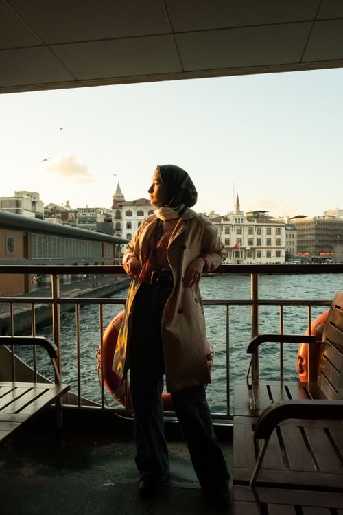 Free Full body of young ethnic female traveler in trendy outfit and traditional hijab leaning on railing of cruise boat and looking away during trip in city at sunset Stock Photo