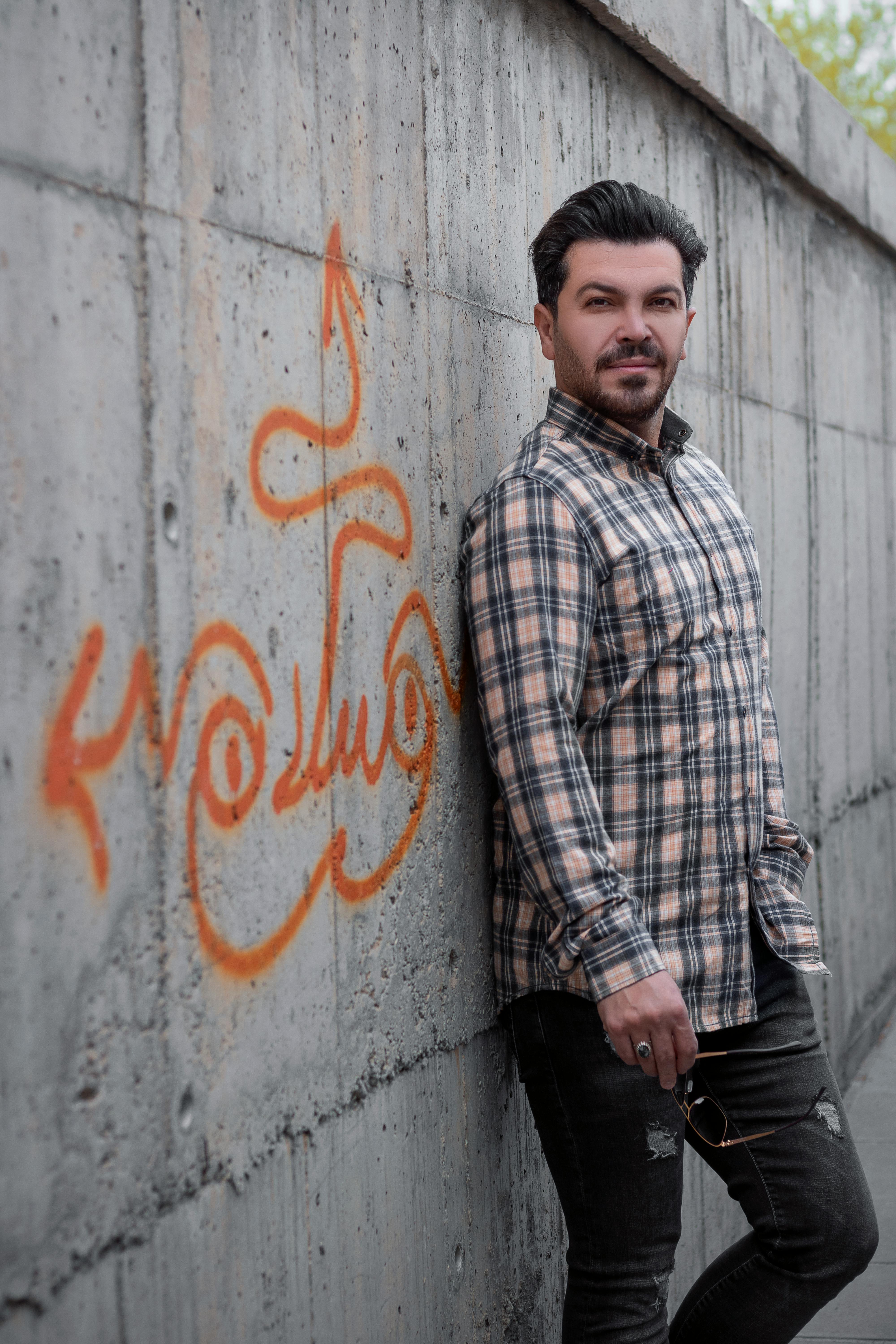 A bearded man in plaid stands against a graffiti-covered wall, exuding casual style.