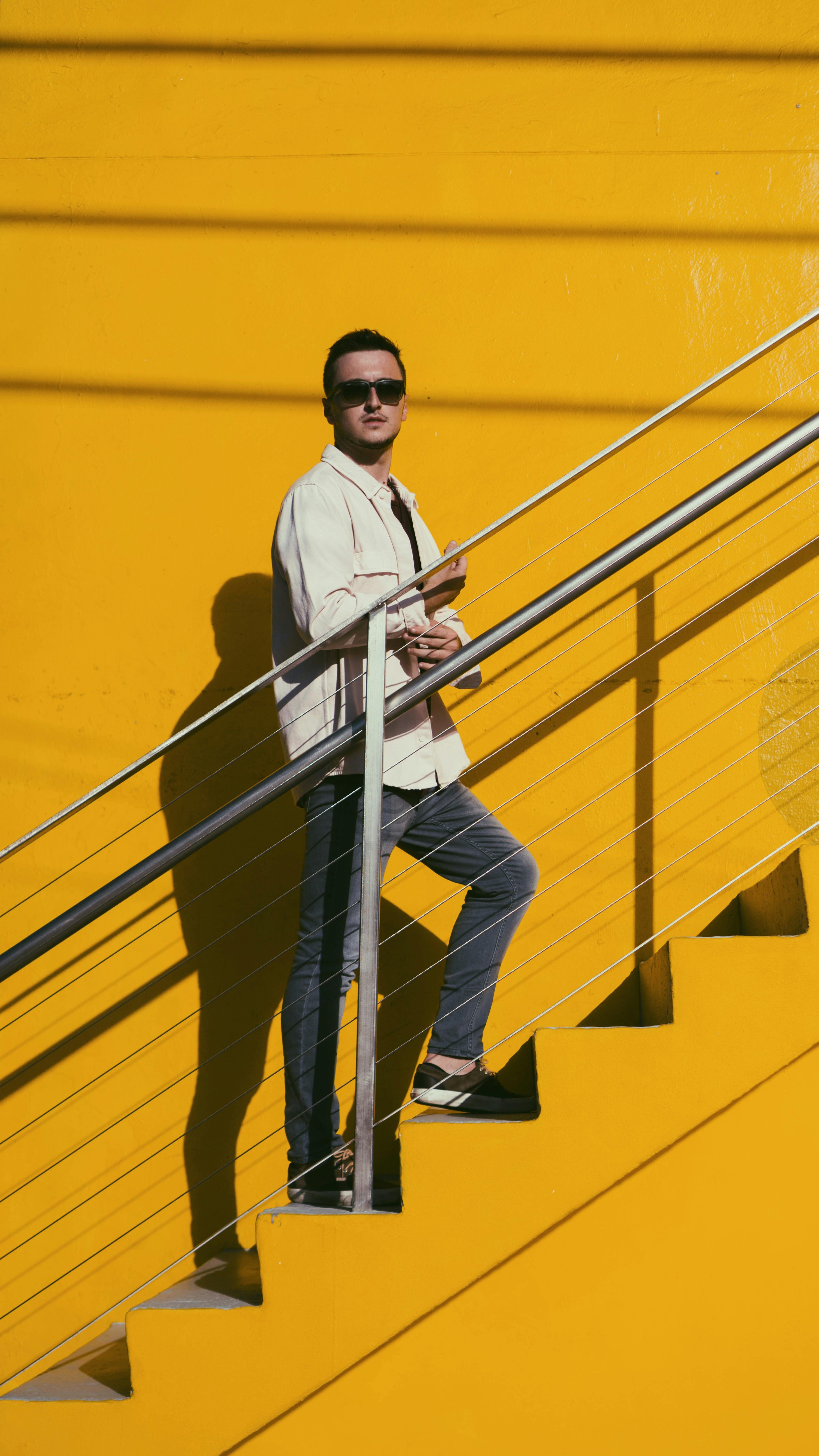 A stylish adult man wearing sunglasses stands on yellow stairs with a striking shadow.