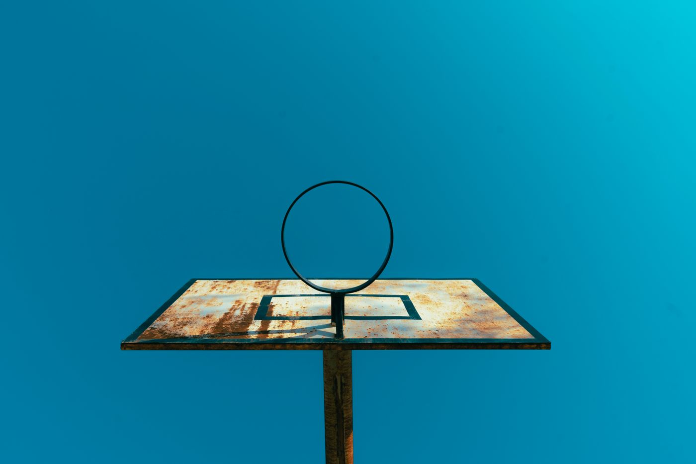 A low-angle shot of a rusty basketball hoop and backboard set against a vibrant blue sky.