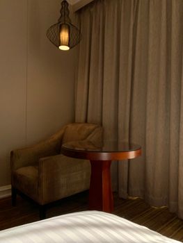 Warm-lit corner of a hotel room featuring an armchair, wooden table, and soft curtains.