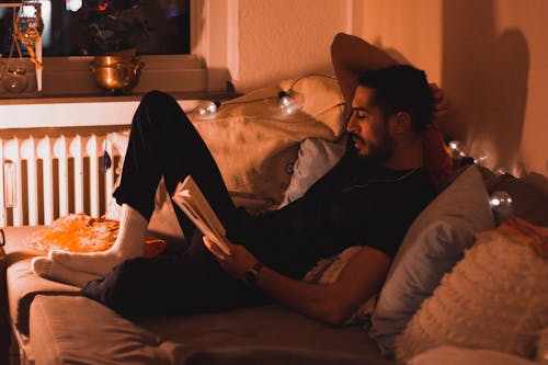 Free Man in Black Shirt and Pants Sitting on Bed Reading Book Stock Photo
