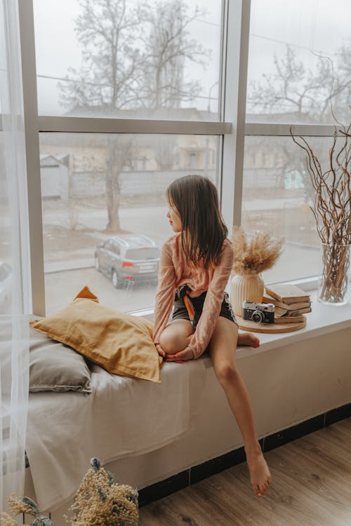 Free Peaceful Asian girl sitting on windowsill in cozy room and looking out of window Stock Photo