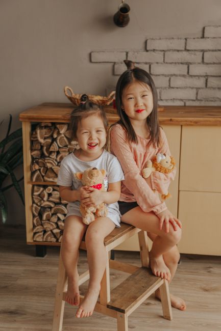 Positive Asian siblings with soft toys sitting on wooden stool at home and looking at camera
