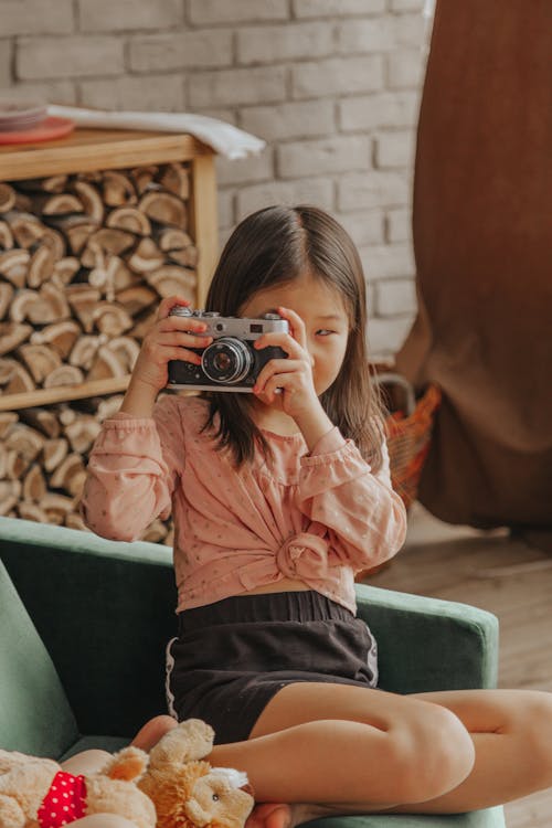Adorable ethnic girl taking pictures on vintage photo camera while sitting on sofa in living room