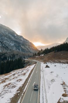 An aerial view of a car driving on a snow-covered road through mountainous scenery at sunset.