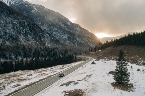 Free A stunning view of a road winding through snowy mountains at sunset in Russia, with pine trees and a vehicle visible. Stock Photo