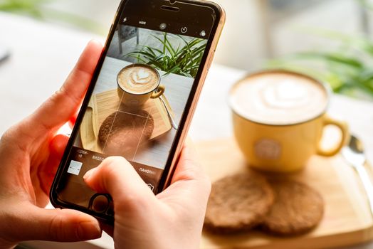 A close-up of latte art being photographed on a smartphone with cookies on a wooden tray.