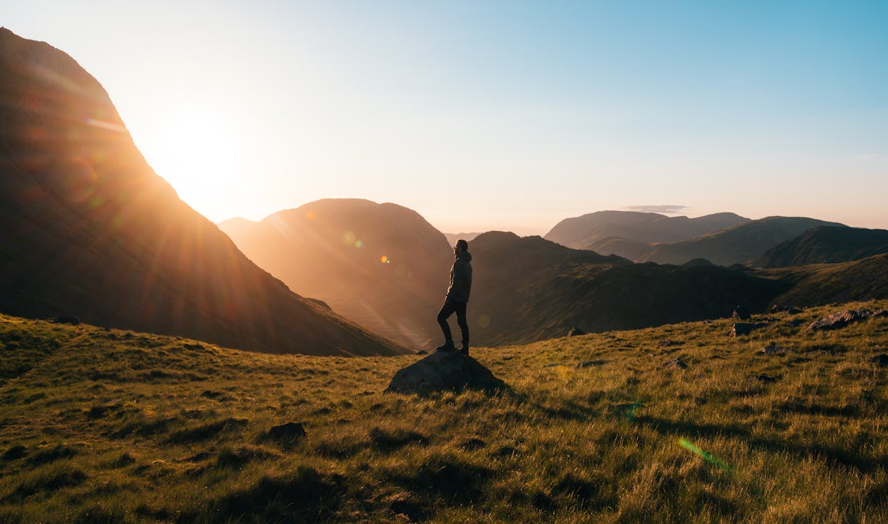 Free Silhouette Photography of Person Standing on Green Grass in Front of Mountains during Golden Hour Stock Photo