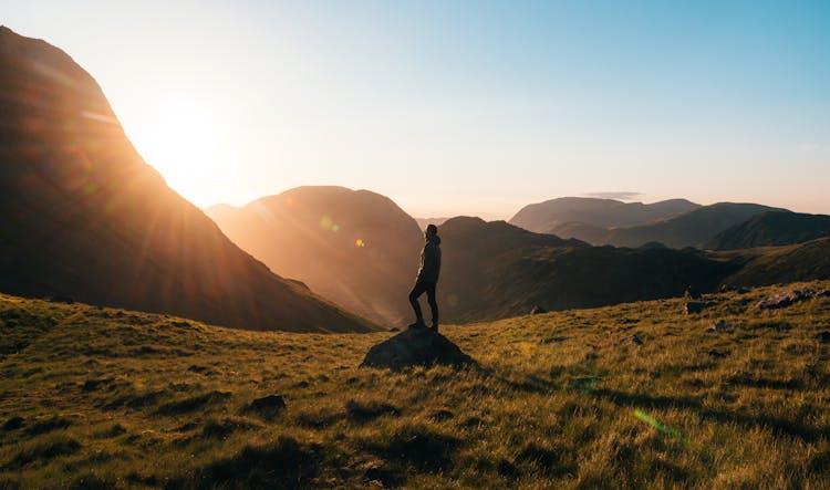 Silhouette Photography Of Person Standing On Green Grass In Front Of Mountains During Golden Hour