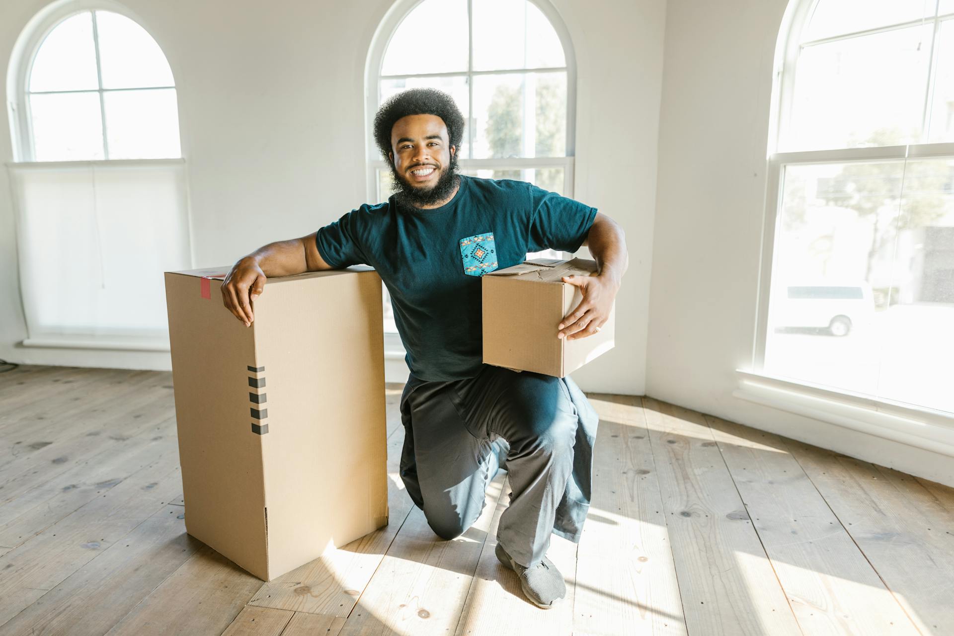 A smiling man with a package and large box in a sunny room. Ideal for delivery and moving concepts.
