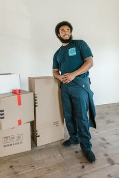 Smiling delivery worker in uniform standing with packaged boxes indoors.