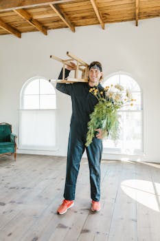 A man in work overalls moves a chair and flowers into a bright new home.