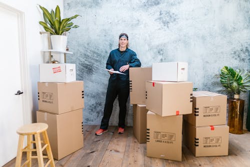 Free Smiling mover in coveralls standing with parcels, clipboard in hand, indoors. Stock Photo