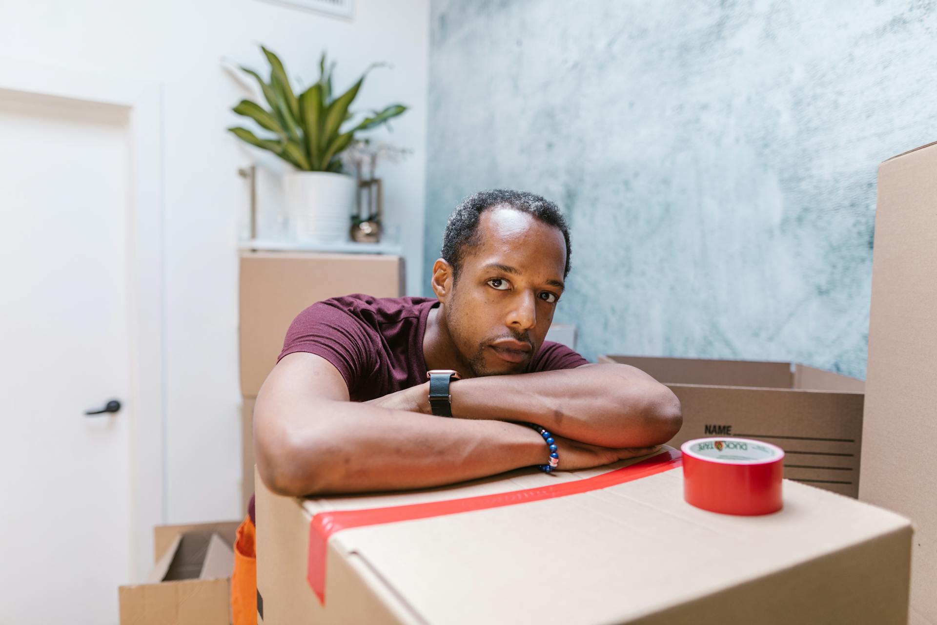 A man resting on cardboard boxes in a room with packing tape visible, suggesting moving or packing activities.