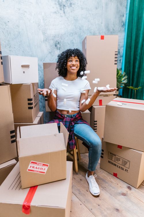 Free A cheerful woman with curly hair sits among packing boxes, tossing foam peanuts. Stock Photo