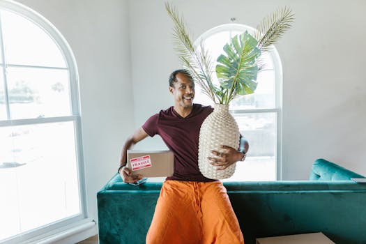 Smiling man holding a fragile box and plant during a home move.